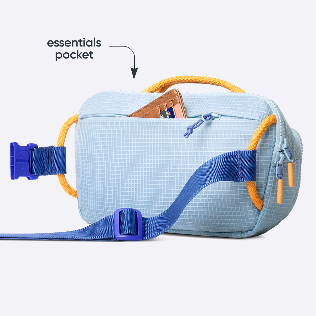 Owala All-Day Crossbody in Rest Azured, light blue ripstop grid belt bag with yellow accent loops and periwinkle strap, shown from the back with a card wallet tucked into the front zip pocket, with text reading "essentials pocket"