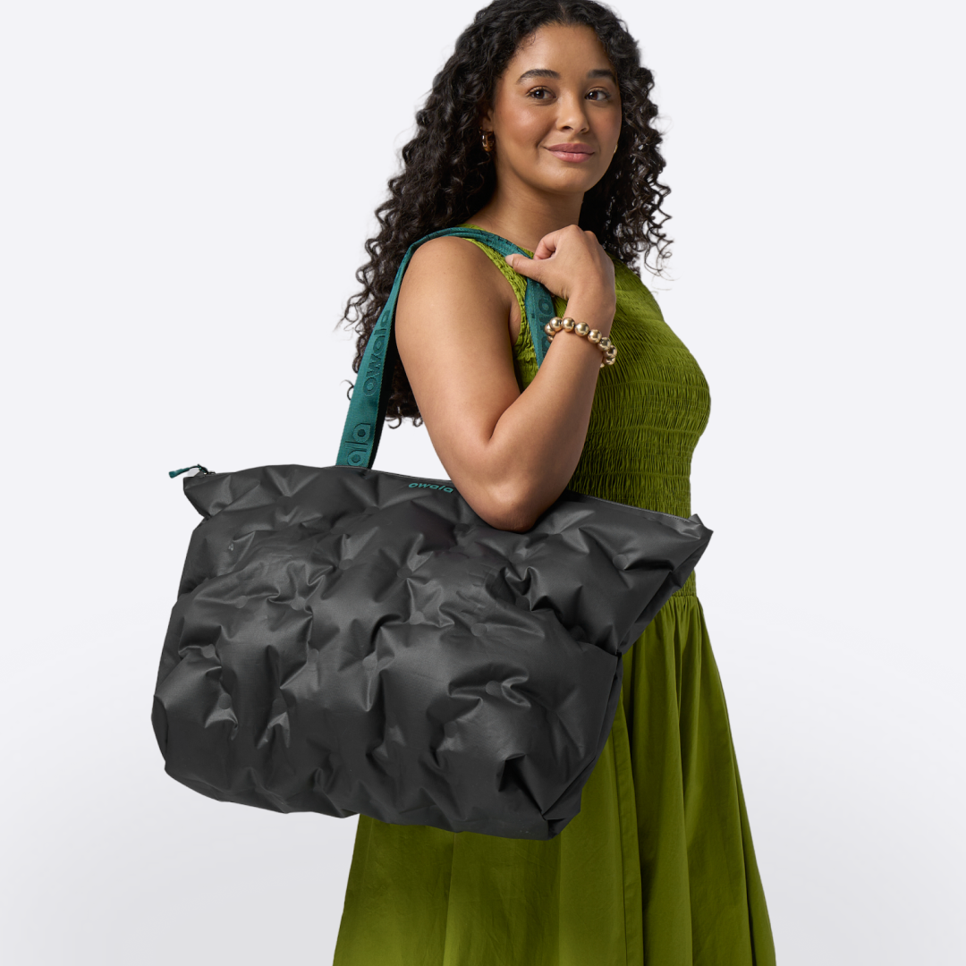 Owala Cloud tote in Night Mode, black inflatable puffer-style bag with teal accent strap carried on the shoulder of a smiling model in a green dress